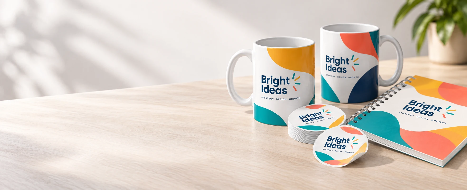 Two branded printed mugs, round logo stickers and a wire bound notebook on a desk